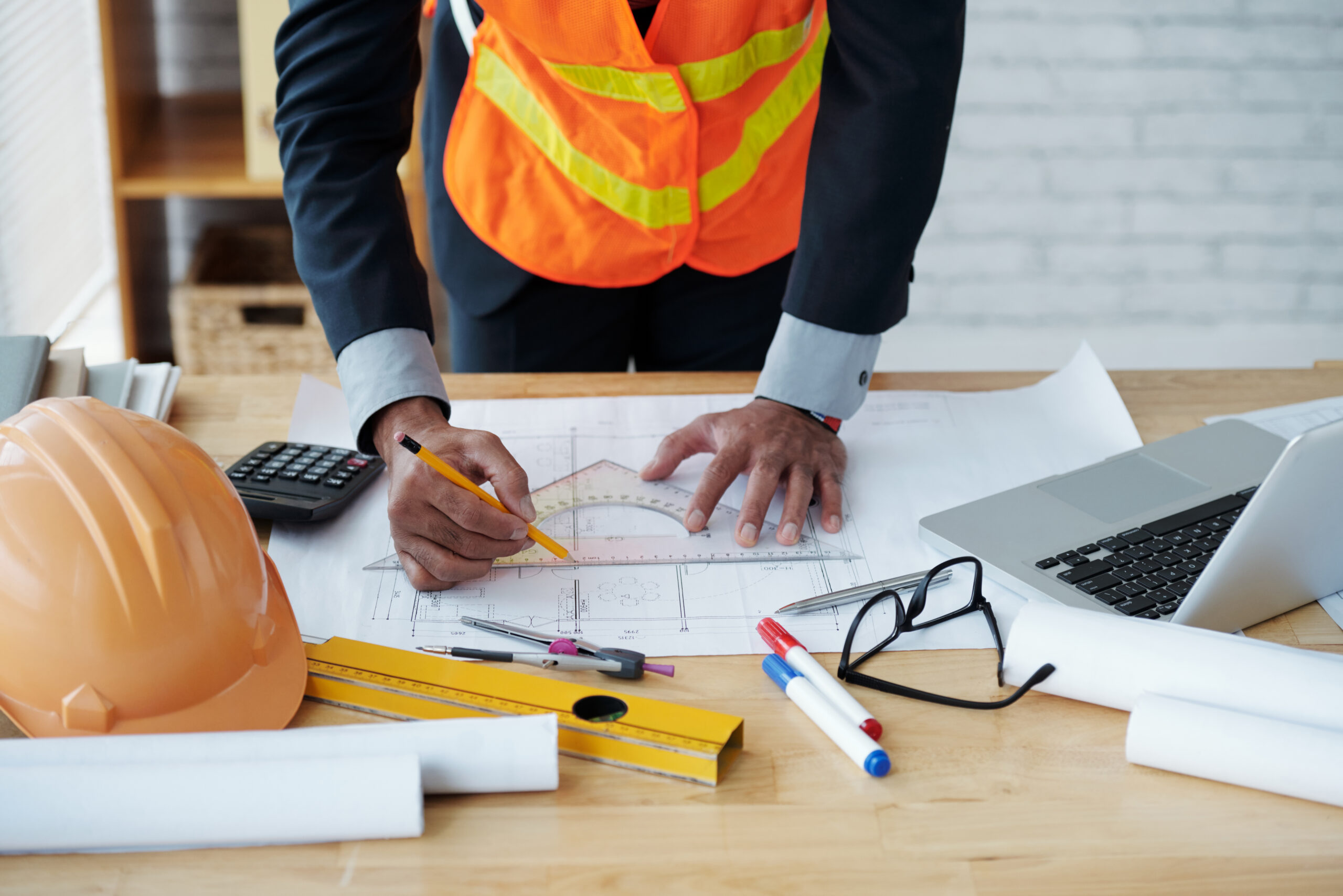 Construction Professional Reviewing Architectural Blueprints Construction professional reviewing architectural blueprints at a desk with a pencil, ruler, hard hat, laptop, and safety vest.